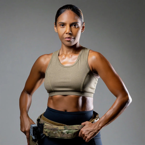A woman posing in a tactical sports bra in olive drab and a camouflage utility belt, set against a solid gray studio background.