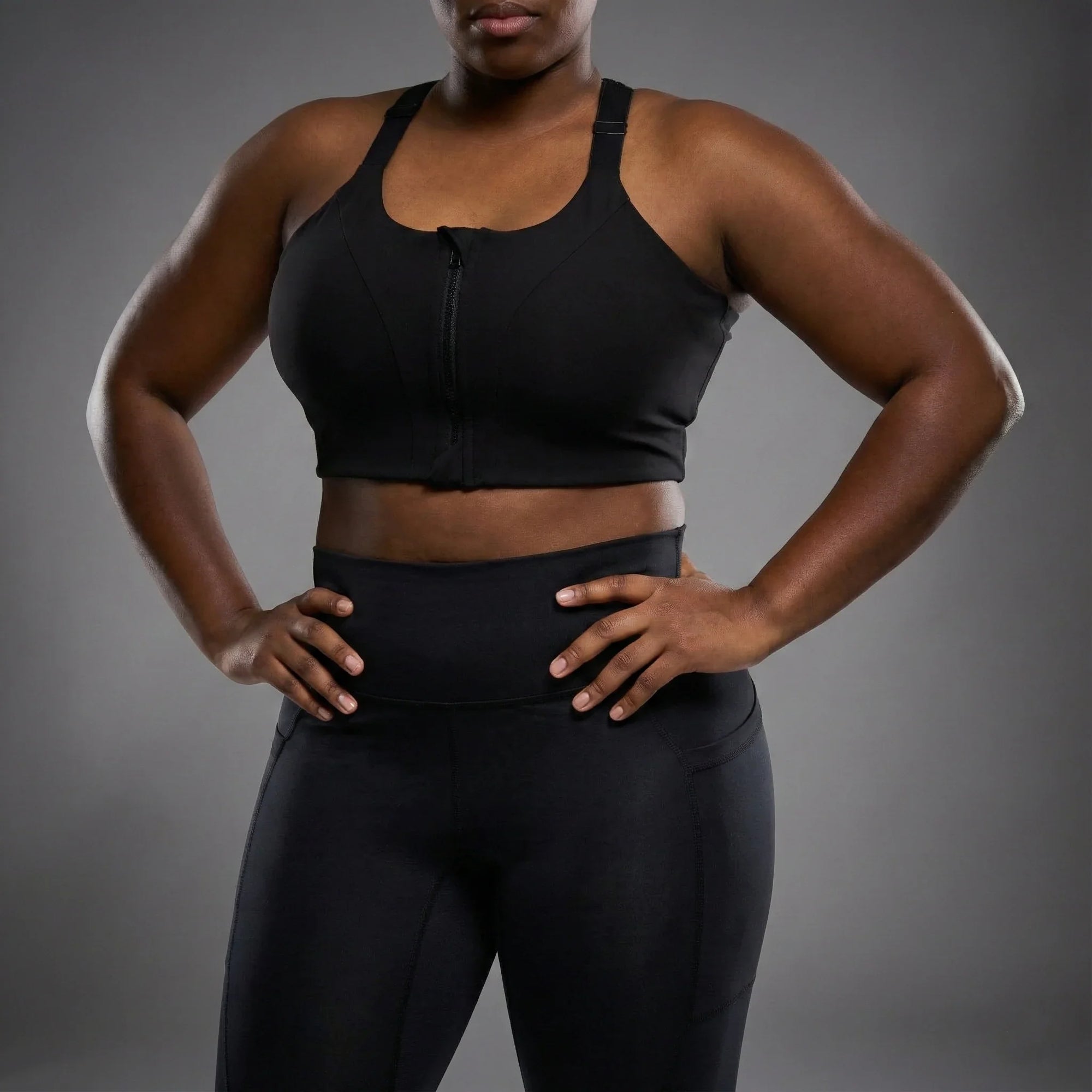 Close-up of a woman wearing black high-waisted leggings and a black zip-front sports bra against gray background