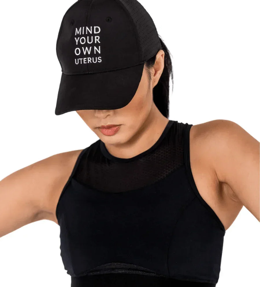 Woman wearing a black cap with embroidered text Mind Your Own Uterus and black athletic top, promoting Oya Femtech Apparel