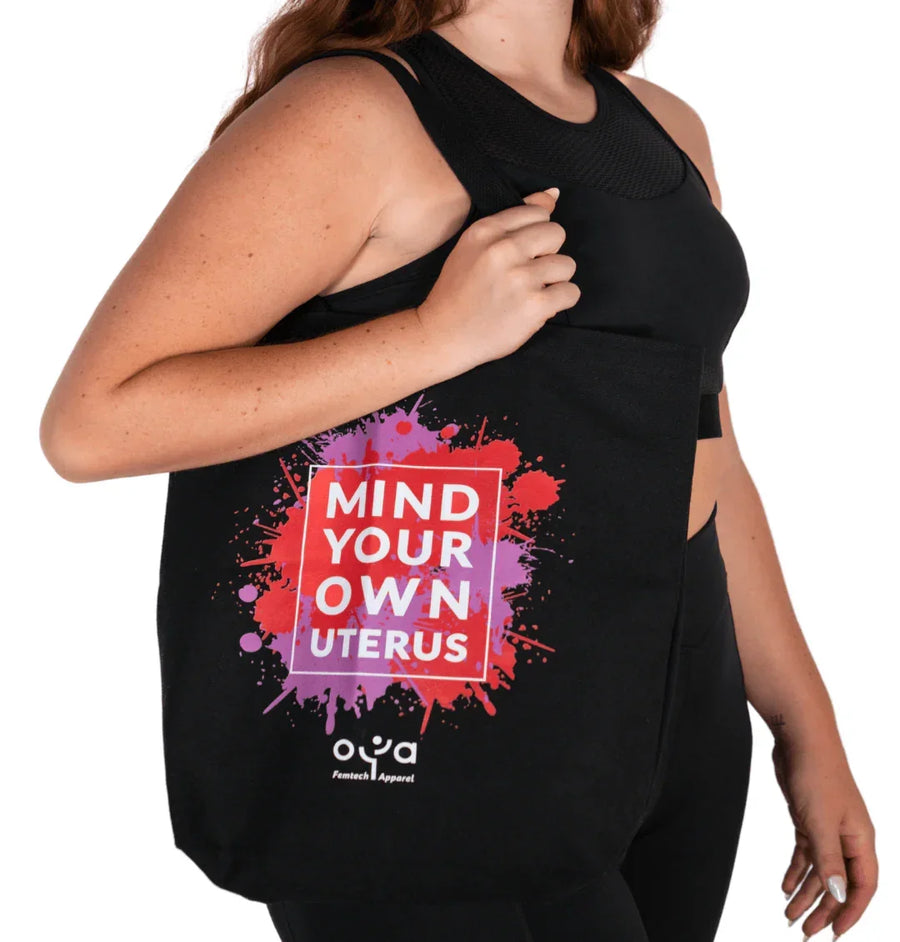 Woman in black activewear holding black tote bag with colorful Mind Your Own Uterus design from Oya Femtech Apparel
